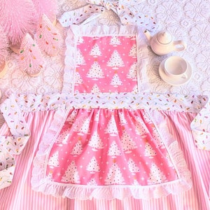 May include: A pink and white striped apron with a pink pocket featuring a white Christmas tree pattern. The apron has a white lace trim and a white ribbon with a bow. The apron is surrounded by pink artificial Christmas trees.