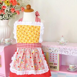 May include: A red and white gingham apron with a yellow floral print and white lace trim. The apron has a red bow at the neck and a white ruffle around the bottom.