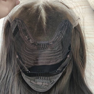 May include: The back of a brown wig with a lace front and adjustable straps. The wig has a brown mesh cap with clips and an elastic band.