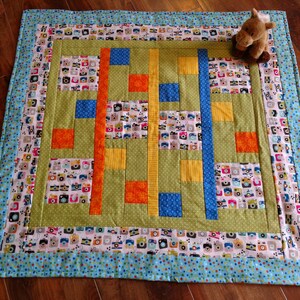 May include: A handmade quilt with a central green panel featuring orange, blue, and yellow vertical stripes and squares. The quilt's border is a light blue with a pattern of cameras and polka dots. A small stuffed animal sits on the quilt.