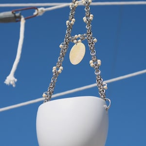 May include: A white ceramic hanging planter suspended by a silver chain with decorative white beads. A small, oval, yellow charm hangs from the chain. The planter is against a bright blue sky.