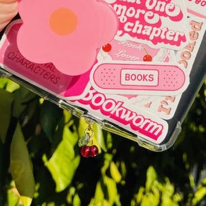 May include: A clear phone case with pink and white stickers. The stickers feature the text "Just one more chapter", "Book Lover", "BOOKS", and "Bookworm". A pink flower-shaped phone grip is attached to the case. A cherry charm hangs from the bottom of the case.