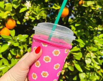 May include: A pink reusable cup sleeve with a white daisy pattern and a smiley face in the centre of each daisy. The sleeve is on a clear plastic cup with a green straw.