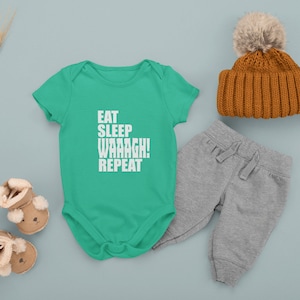 May include: A green baby onesie with the text "EAT SLEEP WAAAGH! REPEAT" in white. The onesie is laid out on a light blue surface with a pair of brown baby booties, a pair of gray sweatpants, and a brown knit beanie with a white pom-pom.