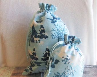 May include: Two drawstring fabric gift bags in varying sizes. The bags feature a light blue background with a winter forest print in dark blue and white. The print includes trees, deer, and snowflakes. A decorative silver branch with white berries is in the foreground.