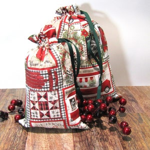 May include: Two drawstring fabric gift bags with a Christmas theme. The bags feature a red, white, and black patchwork design with images of wreaths, trucks, and the words "Hot Cocoa" and "Warm Here". Green ribbon serves as the drawstring.