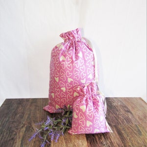 May include: Two pink fabric drawstring bags with a white heart pattern. The larger bag is behind the smaller bag. Both bags have white drawstrings.