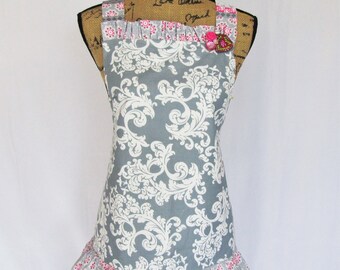 Peut inclure: Un tablier gris et blanc à imprimé damassé avec des touches de rose et des volants. Le tablier comporte une épingle décorative en forme de fleur rose sur la bretelle. Le tablier est présenté sur un mannequin.