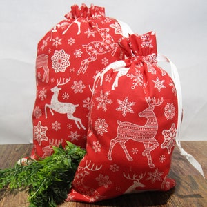 May include: Two red Christmas gift bags with white reindeer and snowflake patterns. The bags have white drawstrings and are made of fabric. One bag is larger than the other, and both are filled.