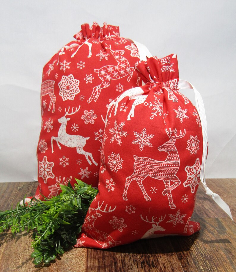 May include: Two red Christmas gift bags with white reindeer and snowflake patterns. The bags have white drawstrings and are made of fabric. One bag is larger than the other, and both are filled.