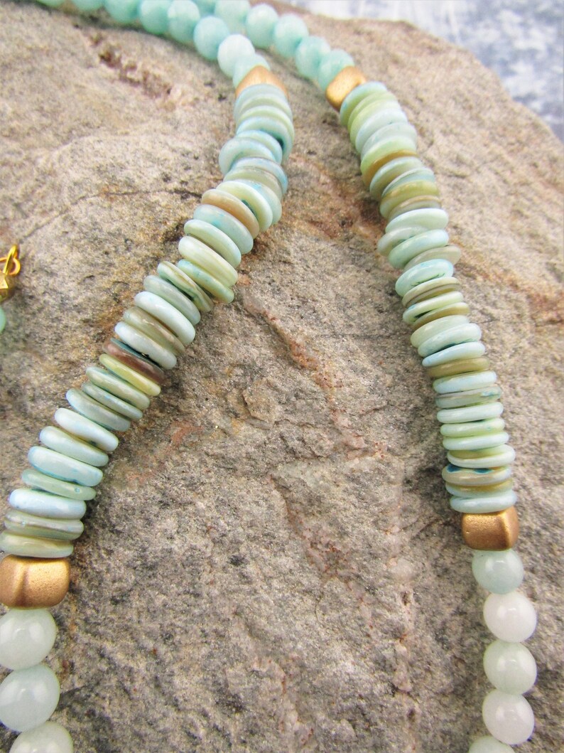 May include: A double-strand necklace made of light blue and green beads with gold accents. The beads are round and flat, and they are strung on a thin gold chain.