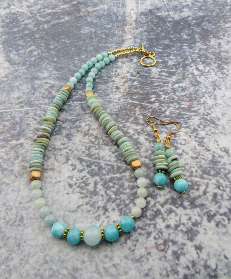 May include: A light blue and green gemstone necklace and earrings set with gold accents. The necklace features a variety of round and flat beads, while the earrings are simple and dangle.