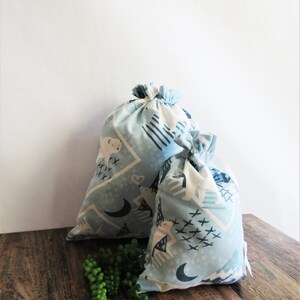 May include: Two light blue drawstring bags with a repeating pattern of white bears, moons, and geometric shapes. The bags are different sizes and sit on a wooden surface. A sprig of green faux succulents is in front of the bags.