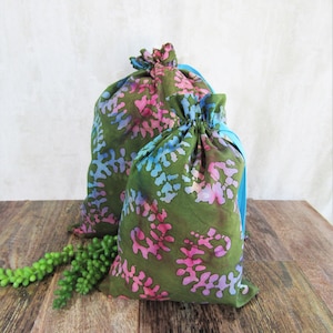 May include: Two drawstring bags with a green, pink, and blue floral pattern. The bags are made of fabric and have a blue drawstring closure. They are sitting on a wooden surface.