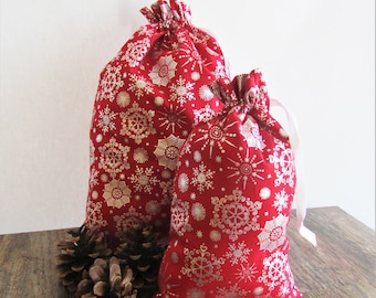 May include: Two red fabric gift bags with a silver snowflake pattern. The bags are different sizes and have a drawstring closure. Several pine cones are next to the bags on a wooden surface.