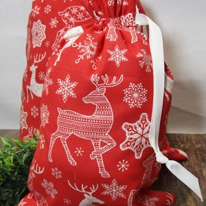 May include: Two red drawstring gift bags with a white reindeer and snowflake pattern. The bags have white ribbon ties and are made of fabric. The bags are on a wooden surface.
