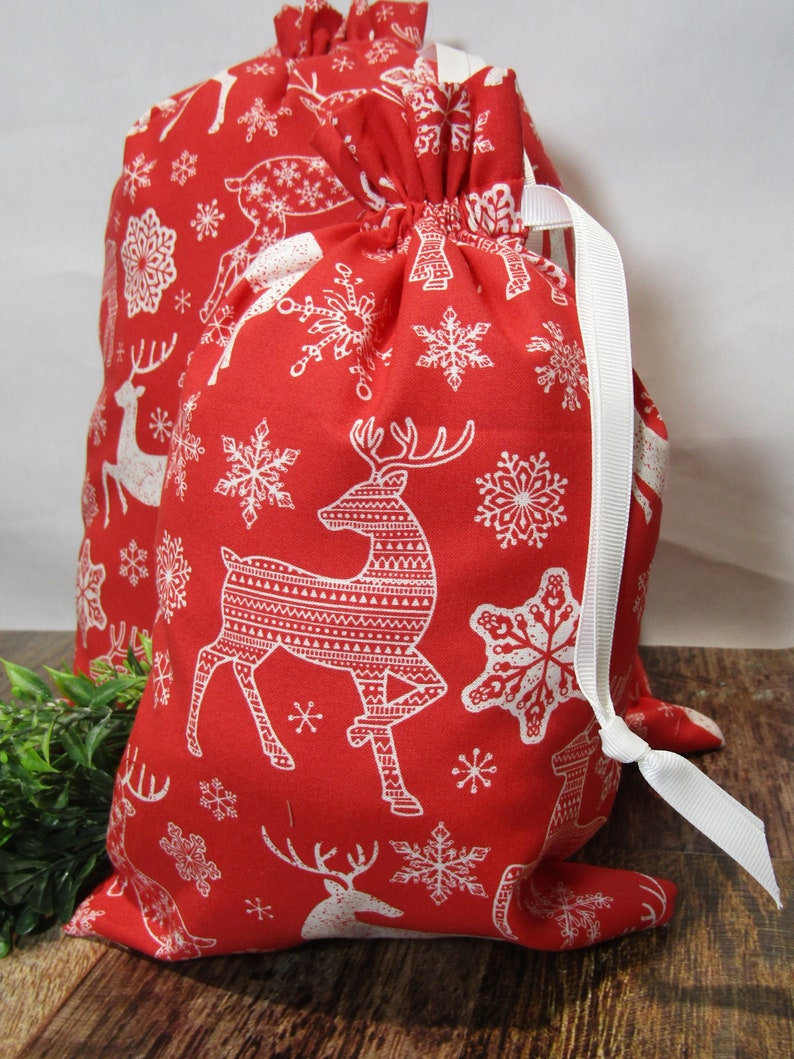 May include: Two red drawstring gift bags with a white reindeer and snowflake pattern. The bags have white ribbon ties and are made of fabric. The bags are on a wooden surface.
