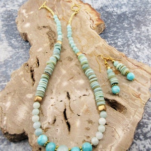 May include: A light blue and green gemstone necklace and earrings set with gold accents. The necklace features round and flat beads, while the earrings have a dangling design.