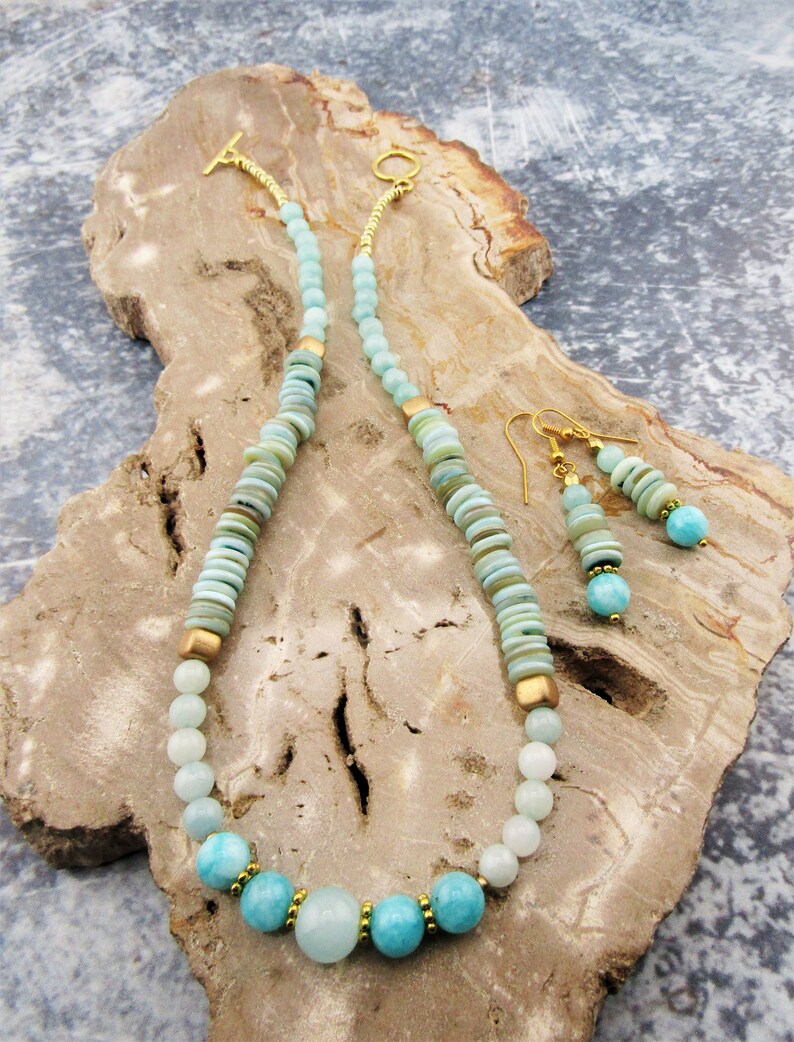 May include: A light blue and green gemstone necklace and earrings set with gold accents. The necklace features round and flat beads, while the earrings have a dangling design.