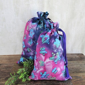Peut inclure: Deux sacs à cordon en tissu avec un motif tie-dye dans des tons de rose, violet et bleu. Les sacs ont des cordons violets.