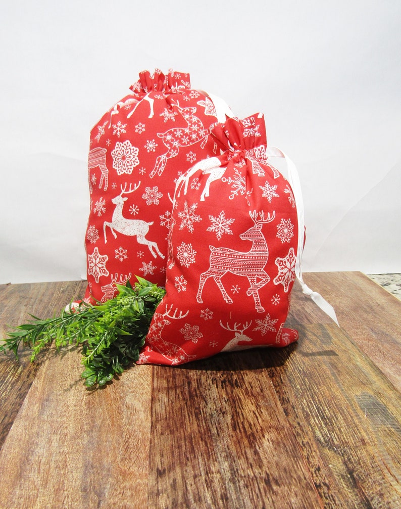 May include: Two red drawstring gift bags with a white reindeer and snowflake pattern. The bags are made of fabric and have white drawstrings. One bag is larger than the other. Greenery is placed in front of the bags on a wooden surface.