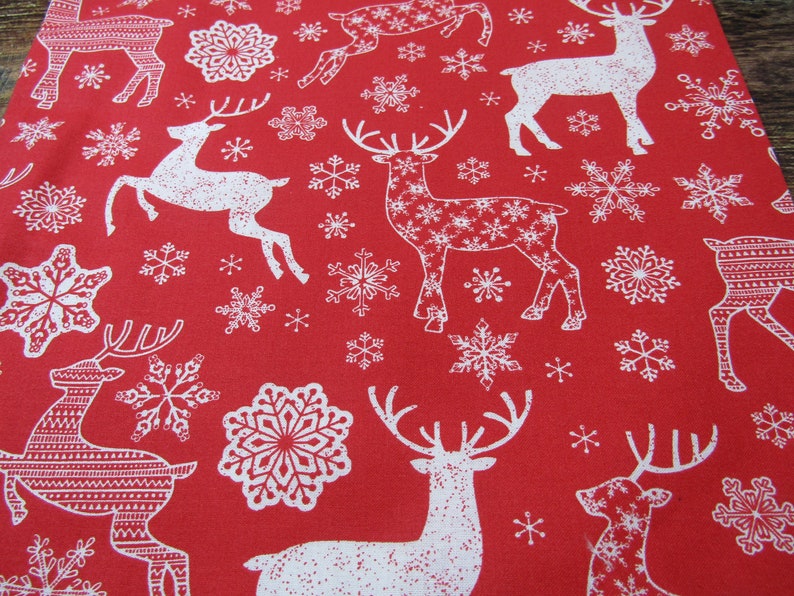 May include: Red fabric with a repeating pattern of white reindeer and snowflakes. The reindeer are in various poses, some with intricate patterns within their silhouettes. The snowflakes vary in size and design, creating a festive holiday theme.