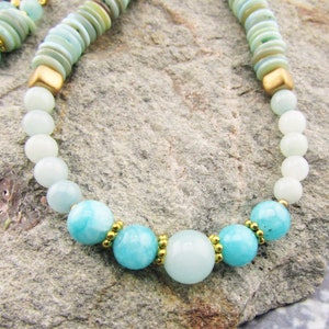 May include: A light blue and white beaded necklace with gold accents. The necklace features round beads and a larger, round, light blue bead in the center.