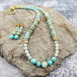 May include: A light blue and green gemstone necklace and matching earrings with gold accents. The necklace features a graduated design with larger, rounder stones at the bottom and smaller, flat stones at the top. The earrings are simple and feature a single, round stone.