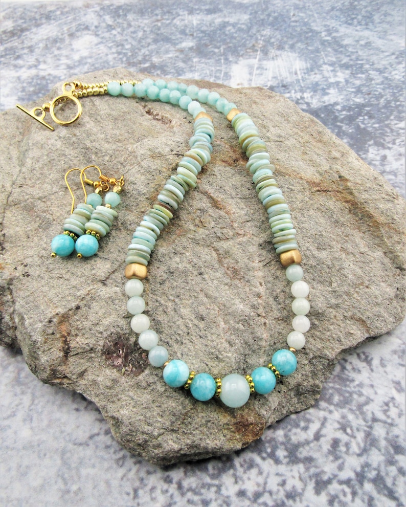 May include: A light blue and green gemstone necklace and matching earrings with gold accents. The necklace features a graduated design with larger, rounder stones at the bottom and smaller, flat stones at the top. The earrings are simple and feature a single, round stone.
