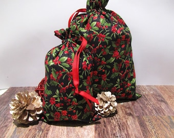 May include: Two drawstring gift bags with a black background, covered in a pattern of red berries and green leaves. The bags have red ribbon ties. Two gold-painted pinecones are in the foreground.