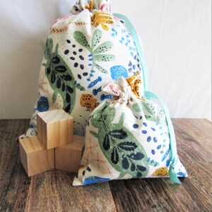 以下が含まれることがあります： 白地に緑、青、黄色の植物や動物のイラストが描かれた、2つの巾着袋。袋は水色のリボンで結ばれています。3つの小さな木製ブロックが袋の前にあります。