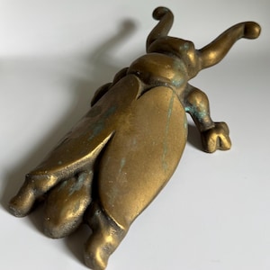 May include: A brass beetle figurine with a weathered, aged patina featuring visible green oxidation in the crevices. The insect-shaped object has two prominent antennae, a segmented body, and two large wing covers extending down the back.