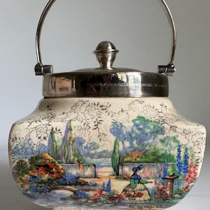 Peut inclure: Pot à biscuits en céramique crème avec une anse et un couvercle argentés. Le pot présente une scène de jardin peinte à la main avec une femme en robe. La base a des pieds décoratifs. Le couvercle est marqué "Yeoman Plate EP Made in England".