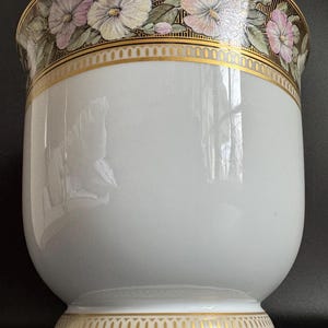 May include: A white ceramic planter with a gold rim and base. The top rim features a floral design with pink, white, and purple flowers. The planter has a wide, rounded body and a slightly flared base, creating a classic, elegant look.