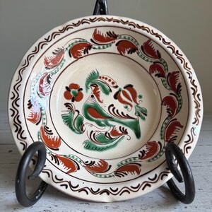 May include: A small white ceramic plate with a brown, green, and orange floral design featuring a bird. The plate has a wavy rim and is on a black metal stand.
