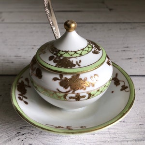 May include: A white porcelain sugar bowl with a lid and a matching saucer. The bowl and saucer are decorated with green and gold floral patterns.