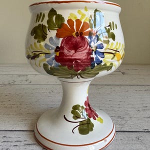 May include: White ceramic pedestal planter with hand-painted floral designs in red, orange, blue, and yellow. The planter has a wide bowl shape and a pedestal base with a decorative rim.