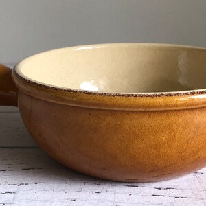 May include: A round, tan ceramic bowl with a handle. The interior is a light cream colour, and the rim has a dark brown trim. The bowl sits on a white wooden surface. This bowl is suitable for serving soups or stews.
