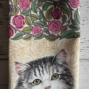 May include: A beige fabric tea towel with a floral pattern of pink roses and green leaves. The bottom half of the towel features a black and white illustration of a cat's face.