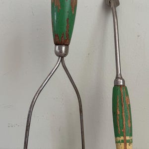 May include: Vintage kitchen tools, including a potato masher and a meat fork. The handles are painted green with distressed details, and the metal components show signs of age. The masher has a looped base, and the fork has two tines.