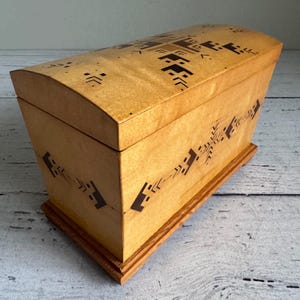 May include: A wooden box with a curved lid and a rectangular base. The box is light brown with dark brown geometric patterns on the lid and sides. The box is sitting on a wooden base.