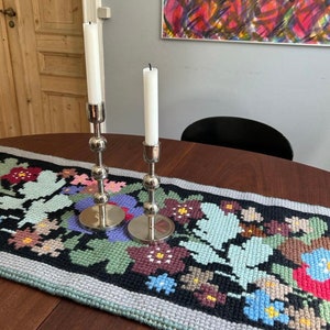 May include: Two silver candle holders with white candles on a table runner with a floral pattern. The runner is black with colorful flowers and a light blue border.