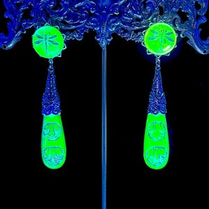 Op de afbeelding: Een paar neon groene oorbellen met een libellenontwerp. De oorbellen hebben een ronde bovenkant met een libel en een druppelvormige onderkant met een decoratief patroon. Het metaalwerk is donkerblauw, tegen een zwarte achtergrond.
