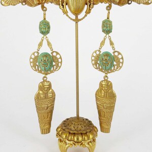 May include: A pair of gold earrings with a scarab beetle, palm tree, and sarcophagus design. The earrings are hanging from a gold stand.