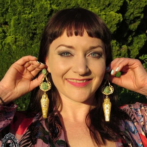 May include: A pair of gold earrings with a green stone and a gold sarcophagus charm. The earrings are dangling and have a detailed design.