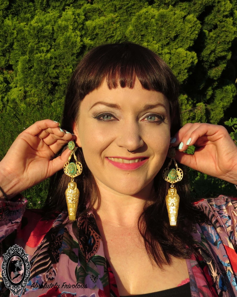 May include: A pair of gold earrings with a green stone and a gold sarcophagus charm. The earrings are dangling and have a detailed design.