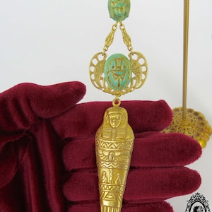 May include: Gold tone pendant necklace with a green scarab beetle charm and a gold tone sarcophagus charm. The necklace is on a red velvet surface.