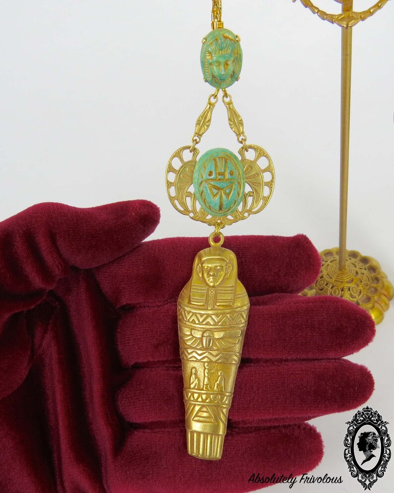 May include: Gold tone pendant necklace with a green scarab beetle charm and a gold tone sarcophagus charm. The necklace is on a red velvet surface.