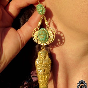 May include: Gold dangle earrings with a turquoise blue scarab beetle and a gold sarcophagus charm. The earrings are hanging from a woman's ear.