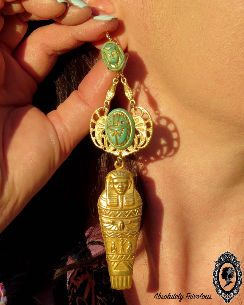 May include: Gold dangle earrings with a turquoise blue scarab beetle and a gold sarcophagus charm. The earrings are hanging from a woman's ear.